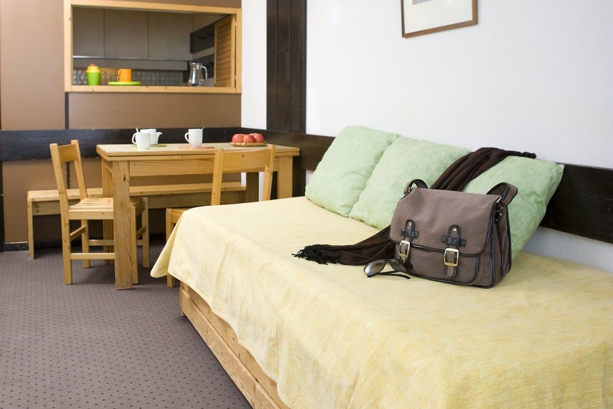 Habitación compacta con cama, escritorio y silla. Incluye una pequeña zona de kitchenette con lavamanos y estante con utensilios de cocina.