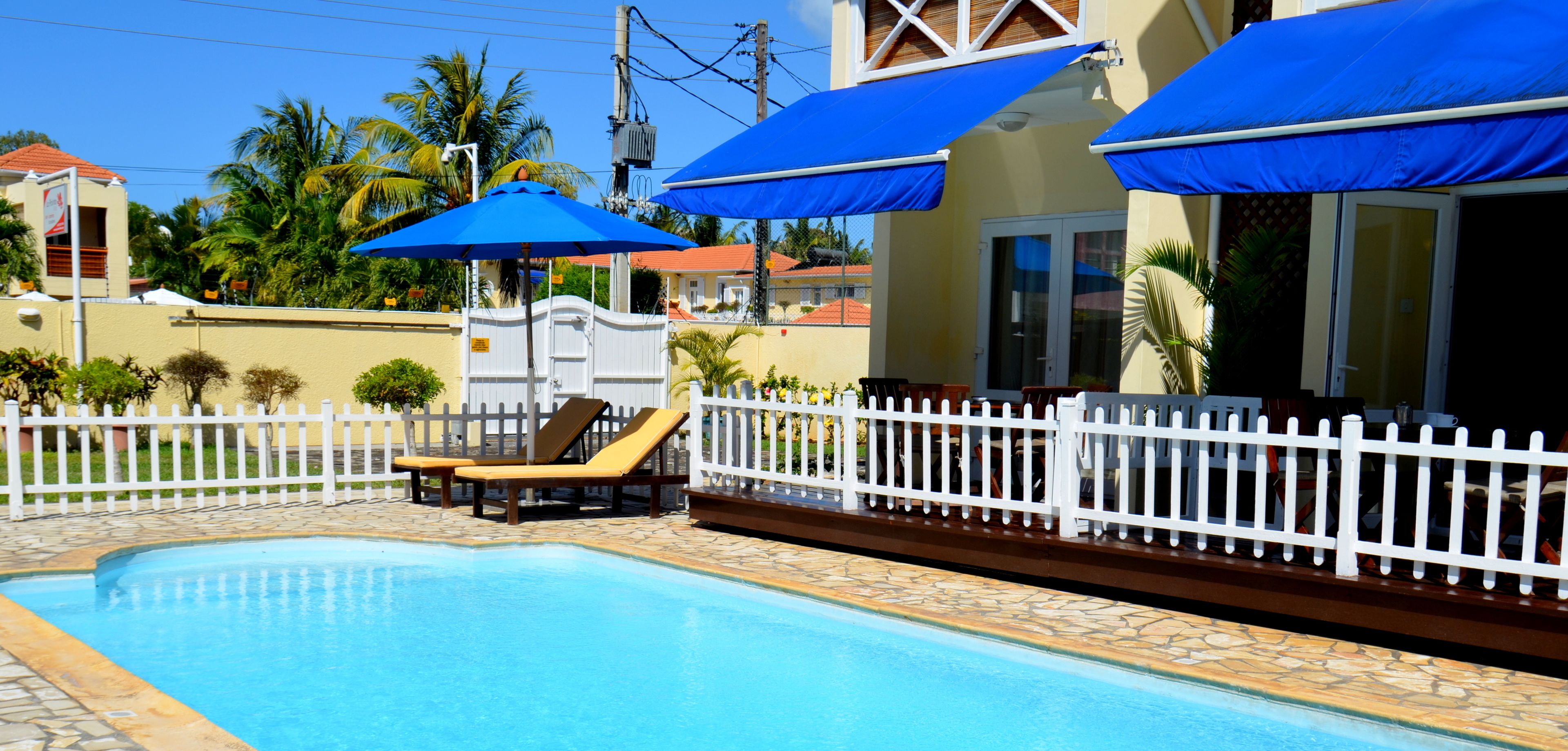 A swimming pool with lounge chairs and blue umbrellas, surrounded by a white fence and palm trees.