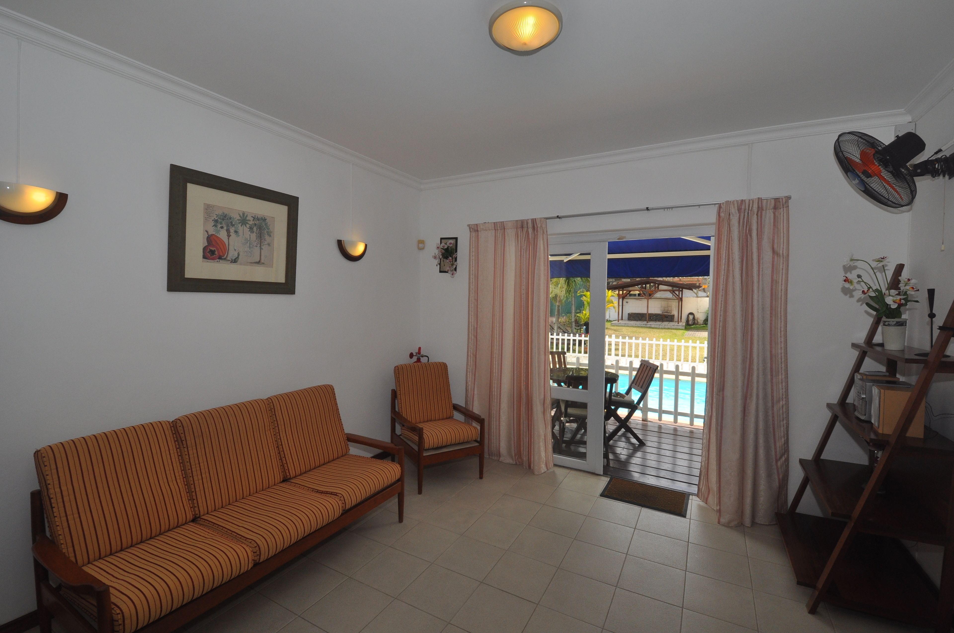 Una sala de estar con sofá, sillas, un ventilador, una estantería con libros y vista a una piscina a través de un ventanal.