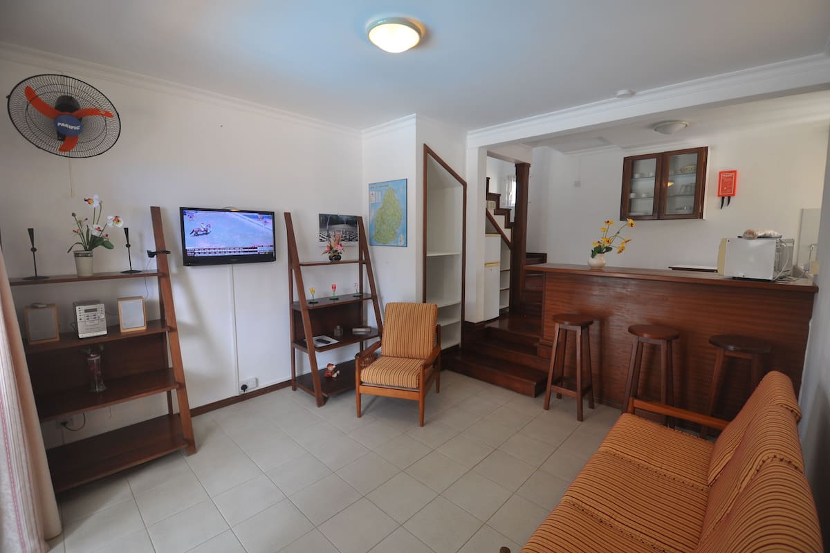 A living room with a TV, wooden furniture, a fan, and a staircase leading to a kitchen.