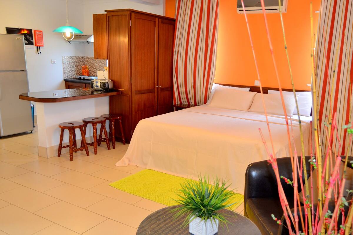 A compact hotel room with a kitchenette, a bed with white bedding, a small dining area with stools, and a potted plant on a round table.