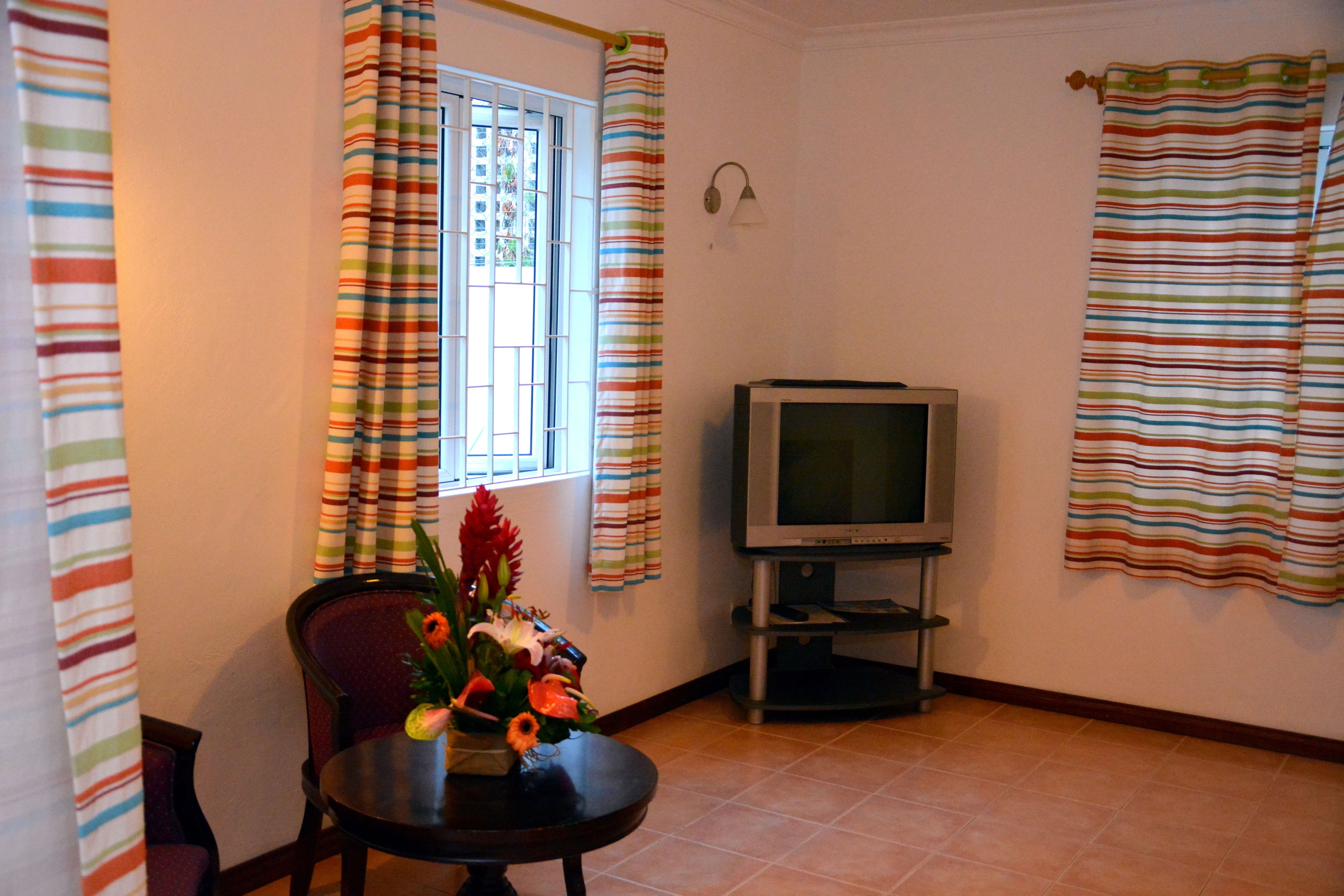 A living room with a television on a stand, a dining table with a vase of flowers, and striped curtains on the windows.