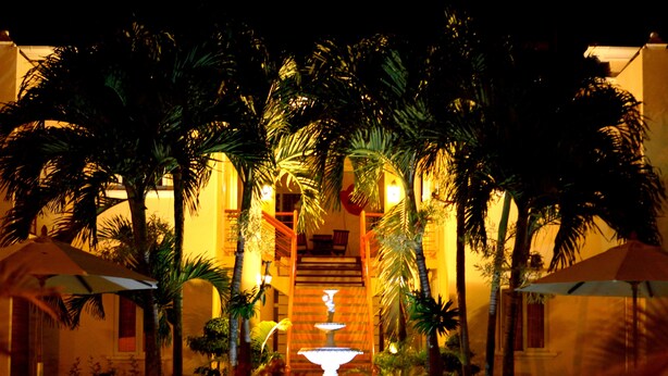 A well-lit outdoor area with a central fountain, surrounded by palm trees and umbrellas, leading to a building entrance. - La Pointe Villas (Grand-Baie)