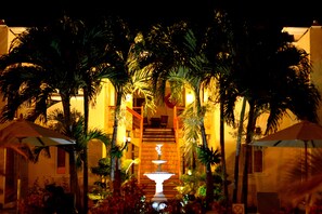 A well-lit outdoor area with a central fountain, surrounded by palm trees and umbrellas, leading to a building entrance.