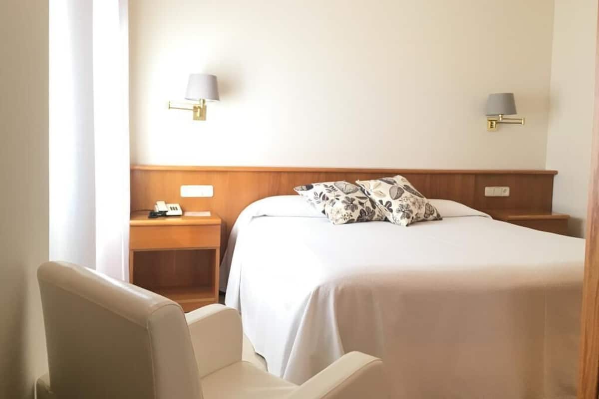 A hotel room with a bed, two bedside tables, a chair, and two wall-mounted lamps.