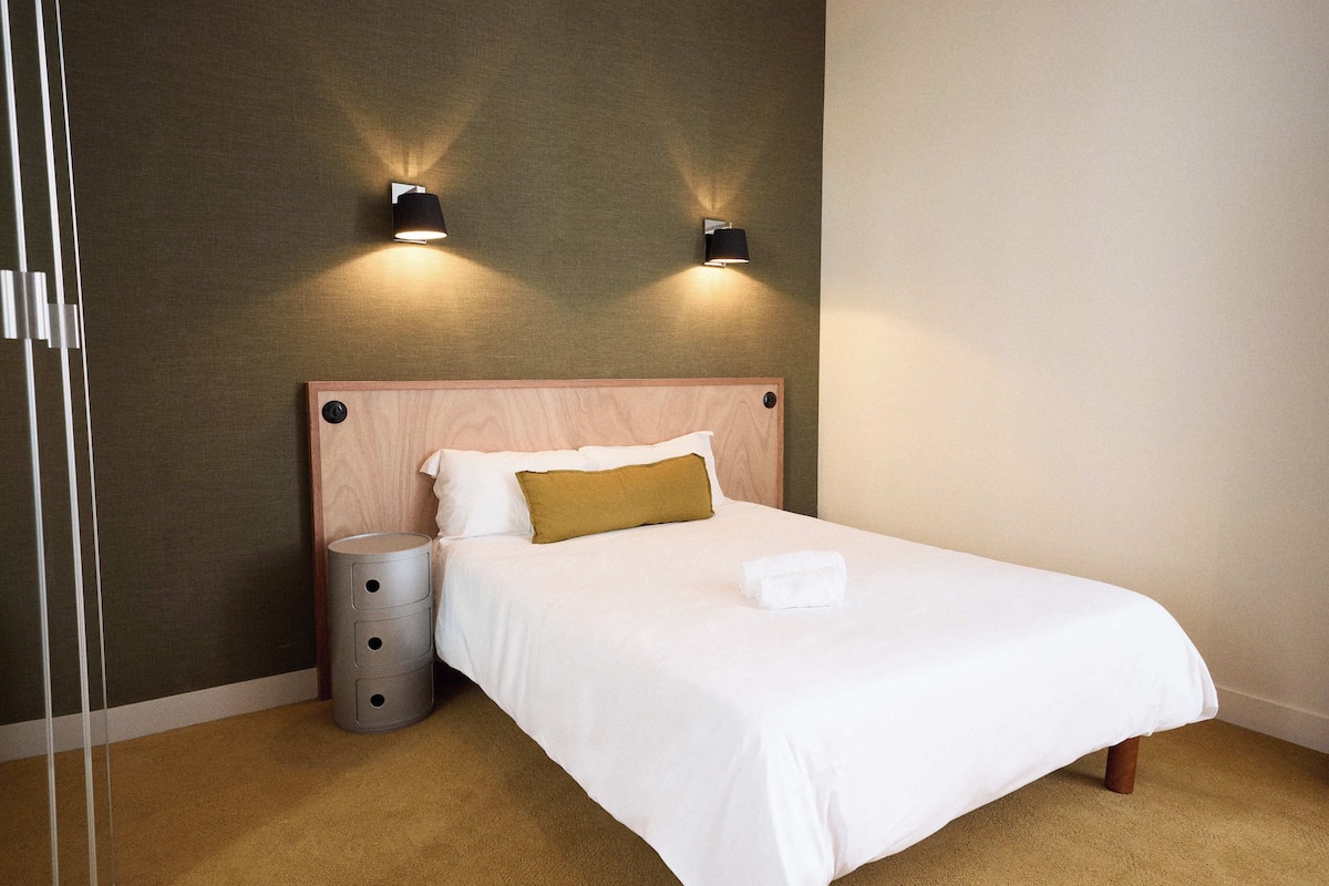 Una cama bien hecha con tablero de madera, dos luces fijadas a la pared, un cubo de basura cilíndrico y piso cubierto de alfombra.