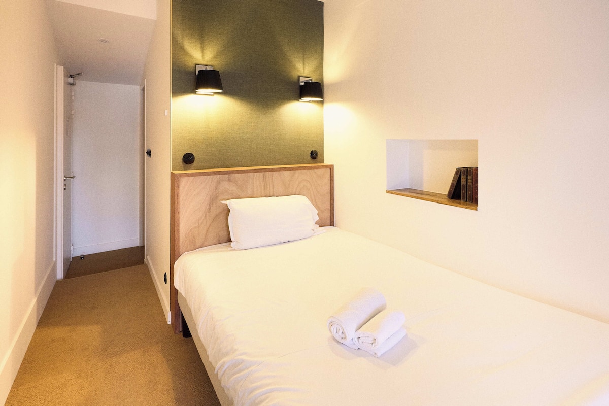 Una habitación de hotel con una cama bien hecha, una mesita de noche pequeña y dos lámparas fijadas a la pared.