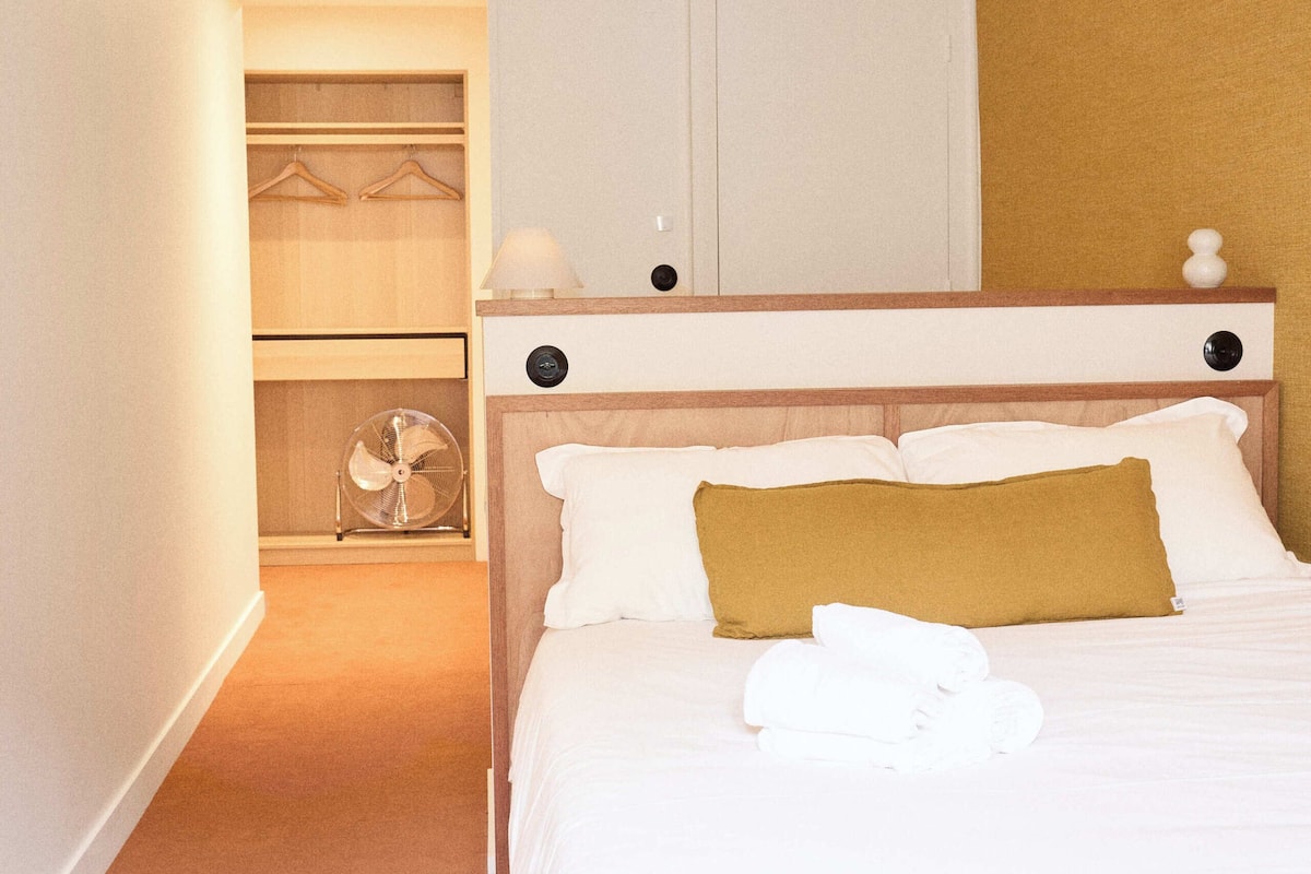 Una habitación de hotel con cama, mesita de noche, lámpara, armario con ropa colgada y ventilador.