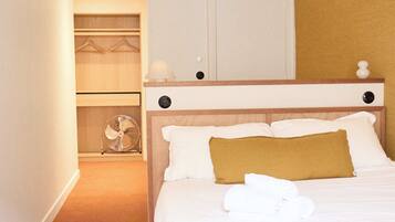 A hotel room with a bed, a bedside table, a lamp, a closet with hanging clothes, and a fan.