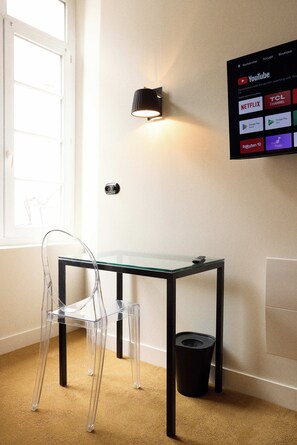 Una habitación con una mesa de vidrio, una silla transparente, un bote de basura negro y una pantalla instalada en la pared que muestra servicios de streaming.
