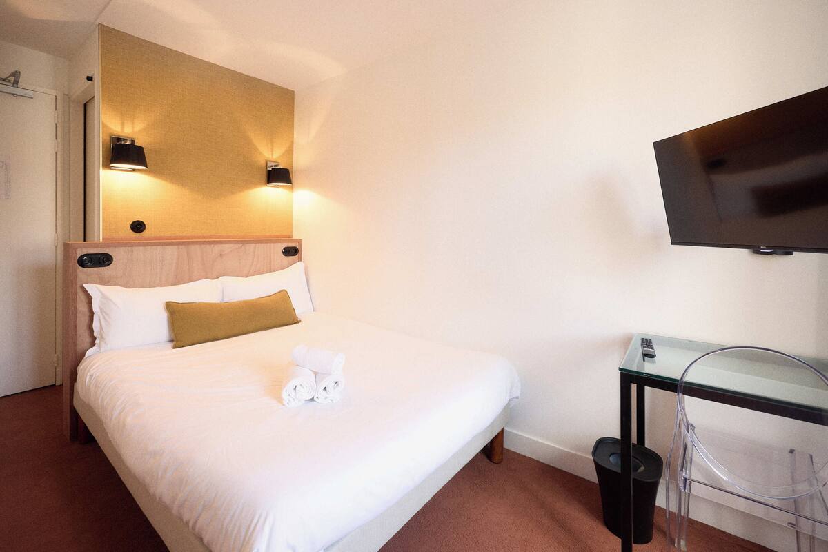 Una habitación de hotel con cama, televisor, mesa de vidrio y un cubo de basura.