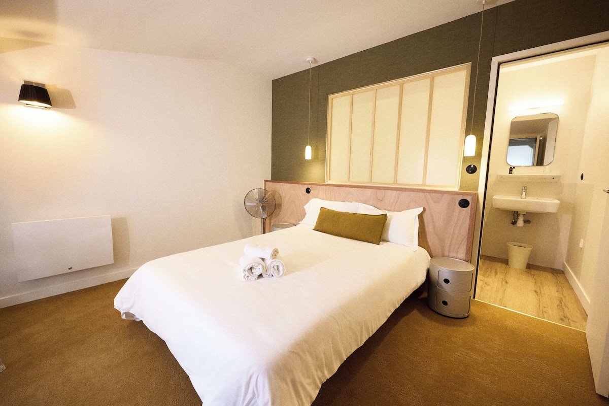 Una habitación de hotel con una cama, un ventilador de pie, un pequeño cubo de basura, un calentador de pared y un espejo sobre el lavabo.
