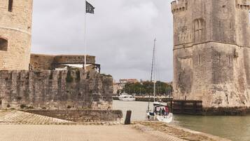 Um castelo de pedra histórico com uma bandeira, um calçadão à beira-mar e um barco atracado nas proximidades.