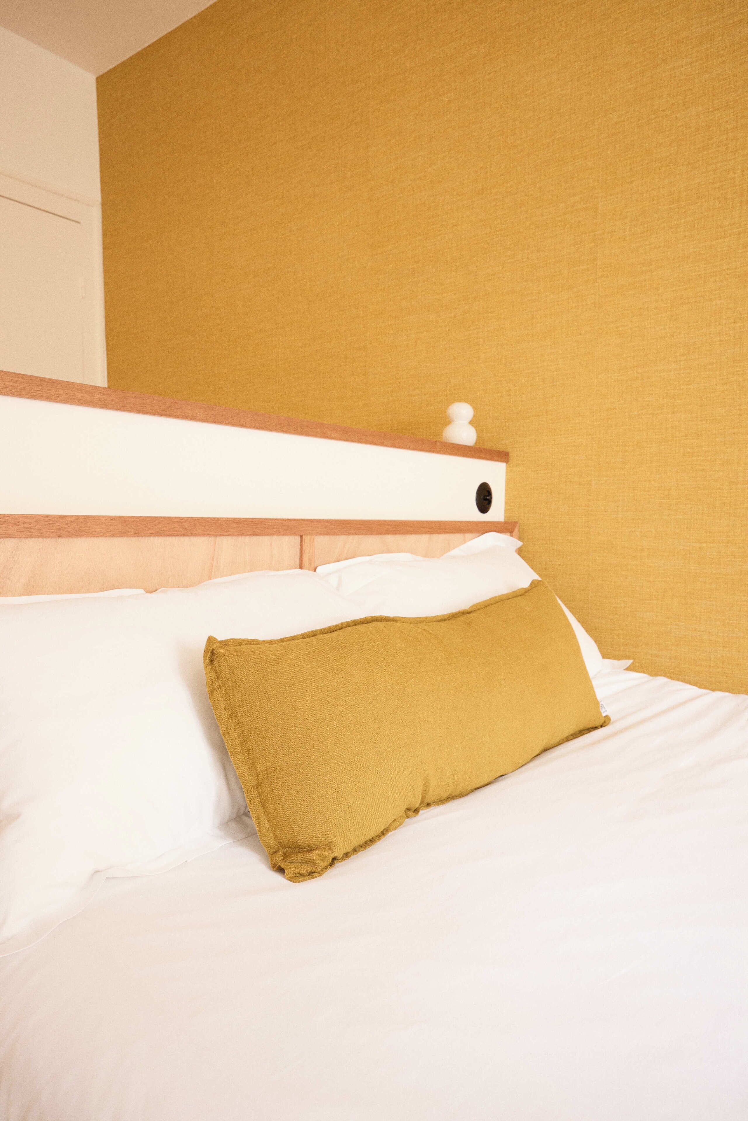 A bed with white linens and a mustard-colored pillow, a wooden headboard, and a plain wall.