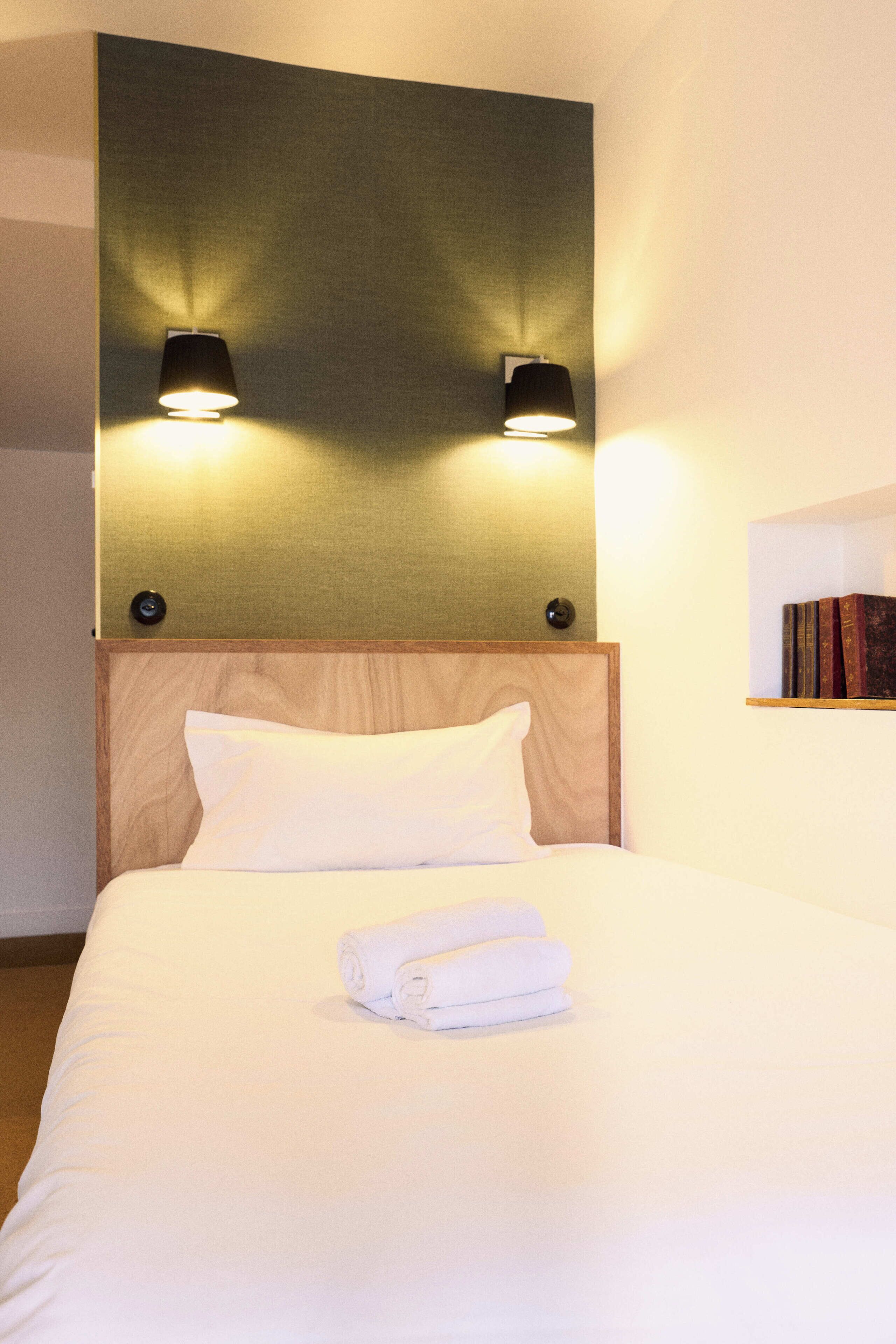 Un lit bien fait, avec du linge de lit blanc et une serviette pliée, une tête de lit en bois et deux lampes fixées au mur.