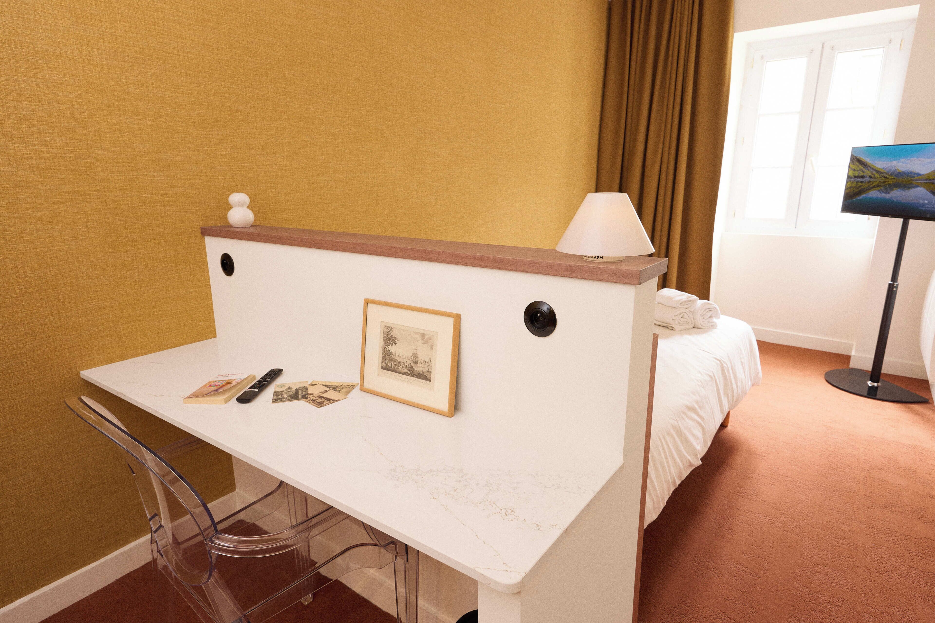 A hotel room with a white desk, a chair, a bed, a lamp, and a painting on the wall.