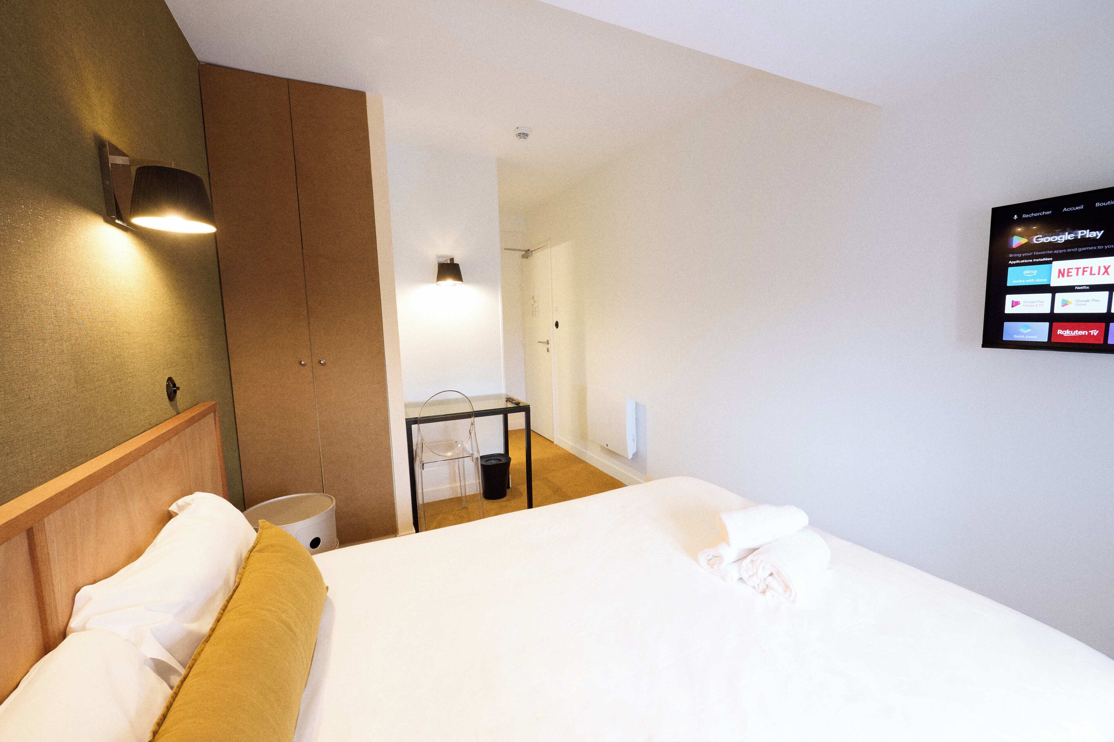 A hotel room with a bed, a wooden headboard, a wall-mounted TV displaying app icons, and a bathroom door.
