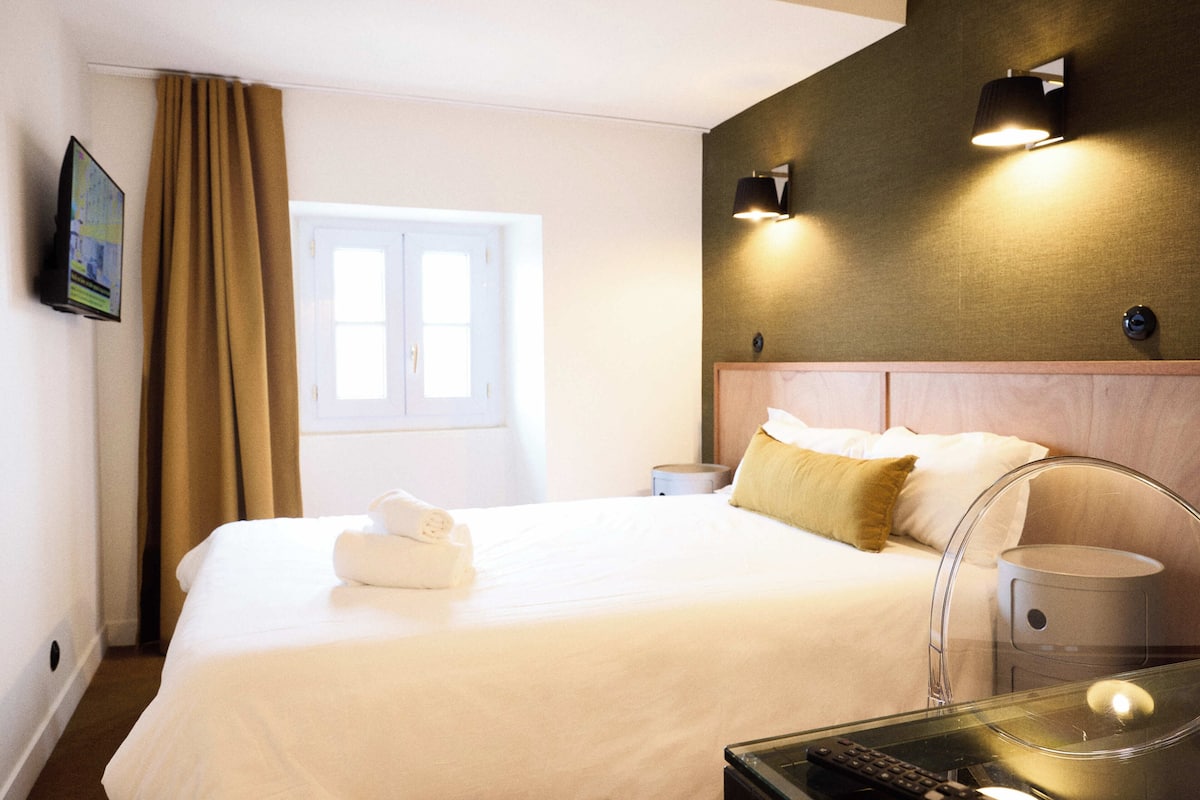 Habitación de hotel con cama, mesita de noche, lámpara, televisor en la pared y ventana con cortinas.