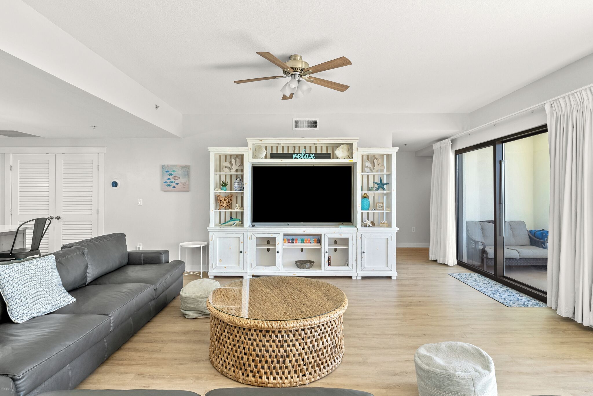 Un salon moderne avec un canapé gris, une table basse ronde en osier, un téléviseur à écran plat posé sur un meuble de télévision blanc et un ventilateur de plafond.