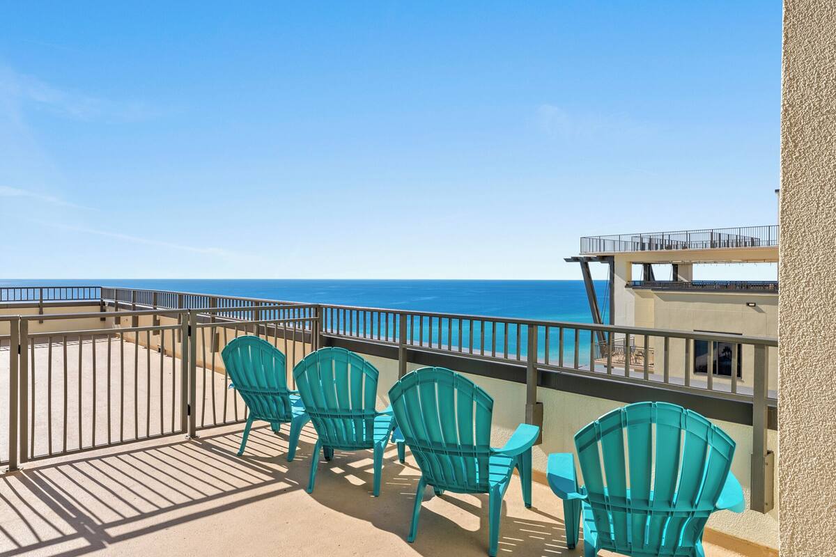 A balcony with turquoise chairs and a table, overlooking a beach and the ocean.