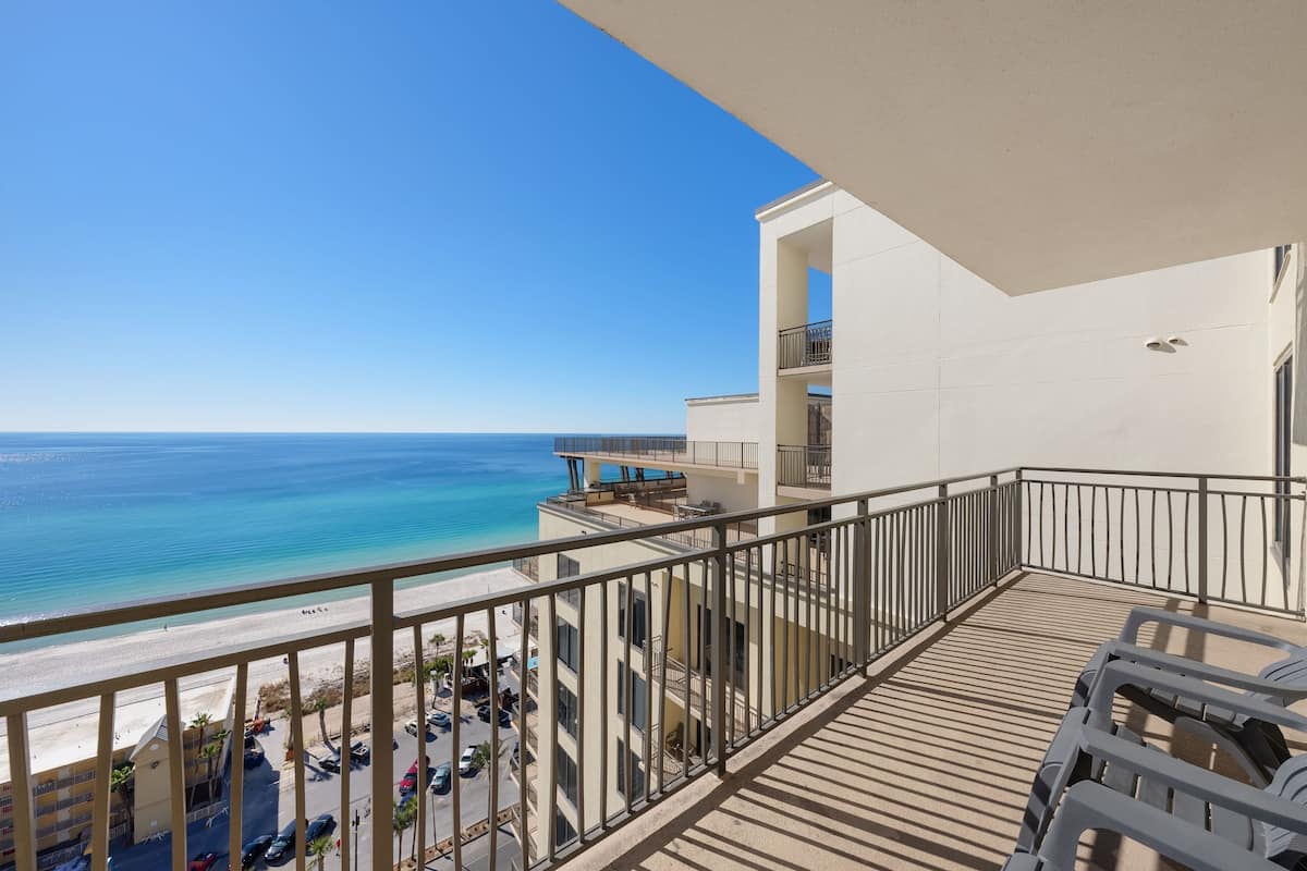 A balcony with a view of the beach and ocean, a pool area, and a parking lot.