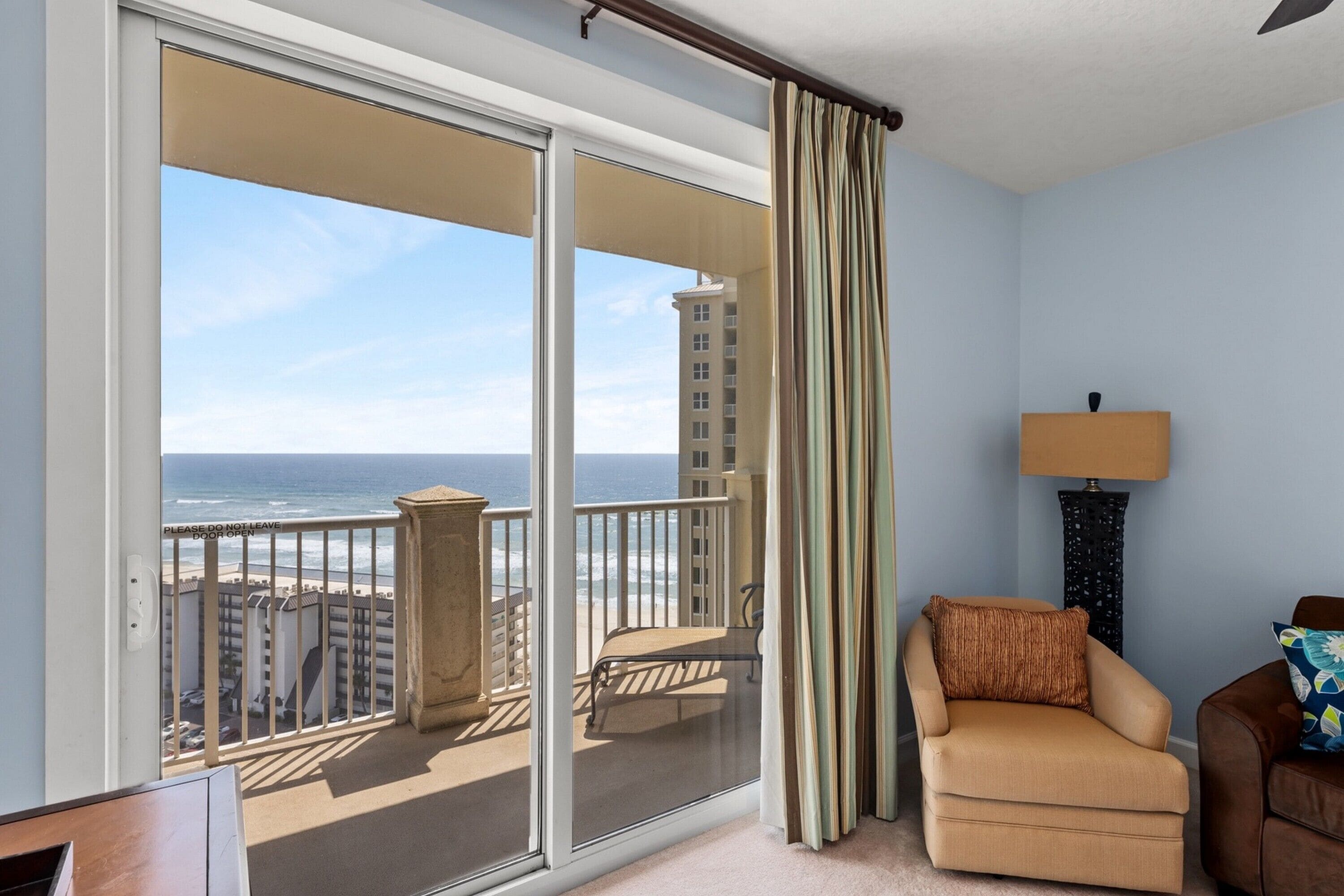 A balcony with a view of the ocean and buildings, a chair, a lamp, and a door with a sign.
