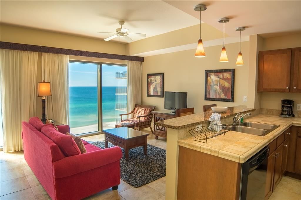 A spacious living room with a red sofa, a coffee table, and a television. There is a kitchen area with a sink and a dining table. The room has a large window with a view of the ocean.