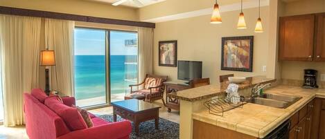 A spacious living room with a red sofa, a coffee table, and a television. There is a kitchen area with a sink and a dining table. The room has a large window with a view of the ocean.
