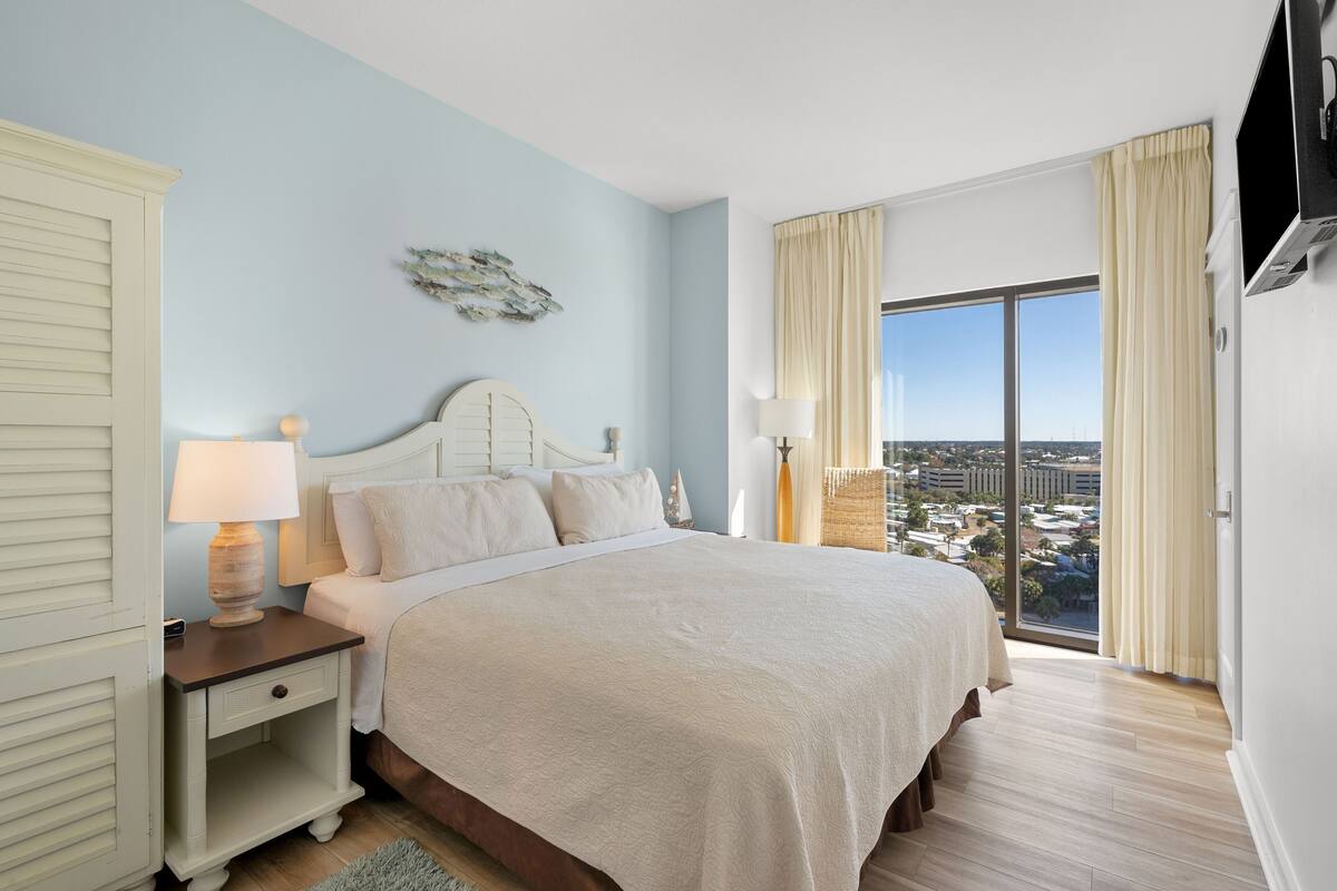 A bedroom with a large bed, a nightstand with a lamp, a view of the city through a large window, and a television mounted on the wall.