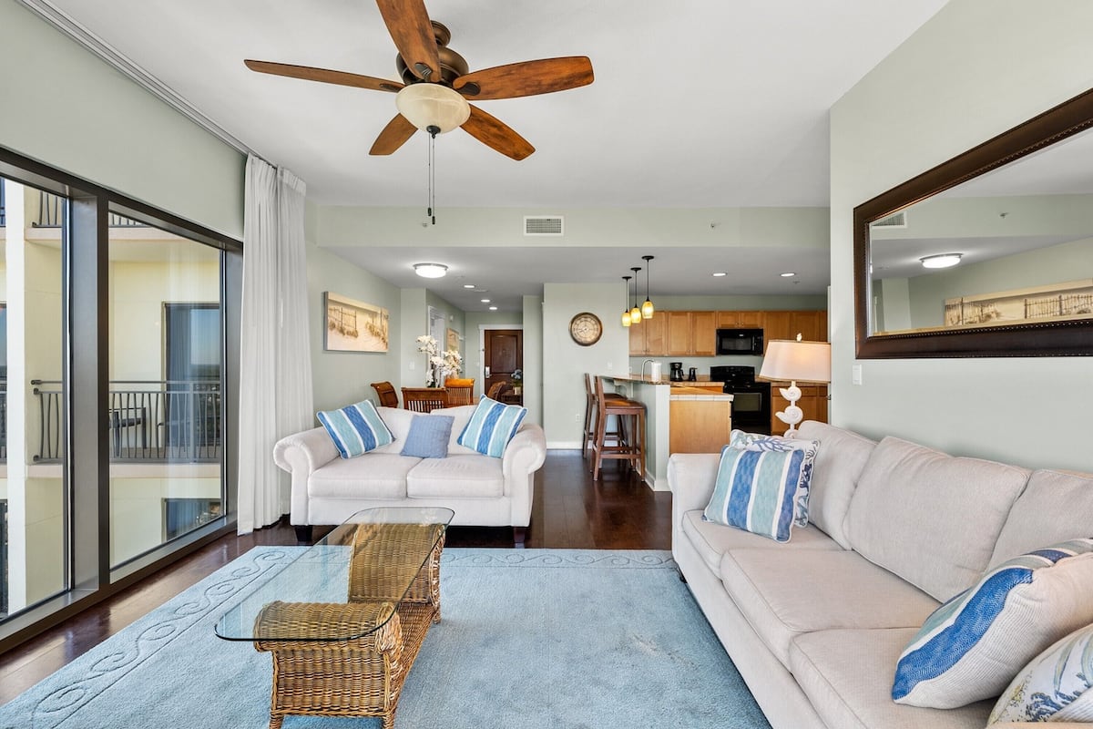 A spacious living room with a ceiling fan, two sofas with blue and white cushions, a wicker coffee table, and a large window offering a view of the balcony.
