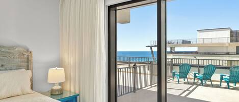 A hotel room with a bed, a nightstand with a lamp, a view of the balcony with chairs, and a clear view of the ocean.