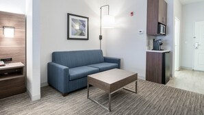 A modern hotel room with a blue sofa, a wooden coffee table, a framed abstract painting, a desk with a telephone, and a kitchenette with a microwave.