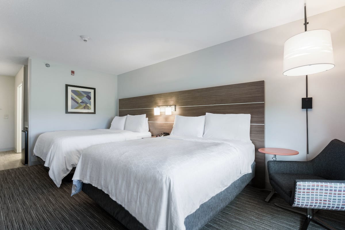 A hotel room with two beds, a small table, a chair, and a wall-mounted lamp.