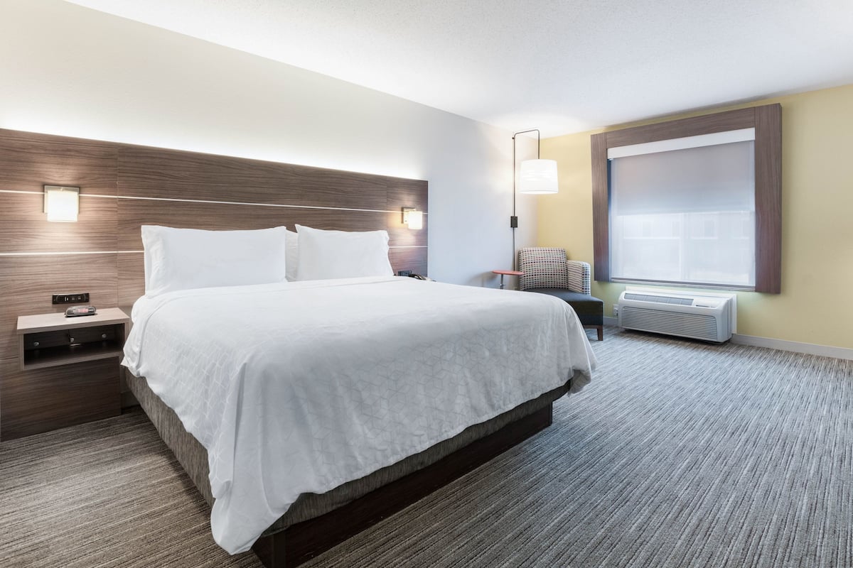 A hotel room with a large bed, a nightstand, a chair, and a window with blinds.