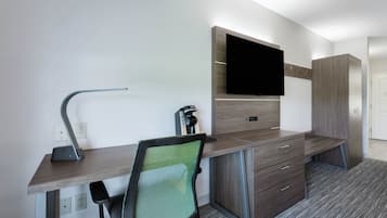 A modern hotel room with a wooden desk, a green mesh office chair, a flat-screen TV mounted above the desk, and a wardrobe to the right.
