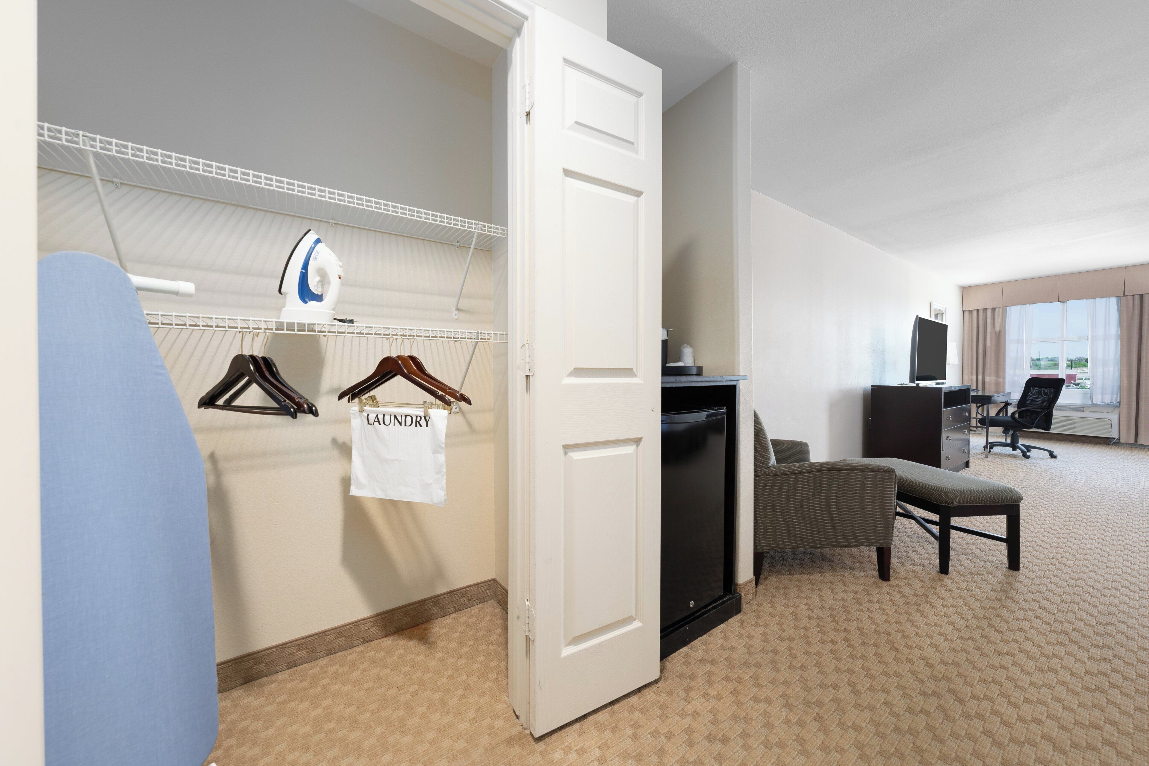 Une chambre d’hôtel comprenant un lit, un bureau, une chaise, une armoire et un espace pour faire la lessive avec un portant à vêtements.