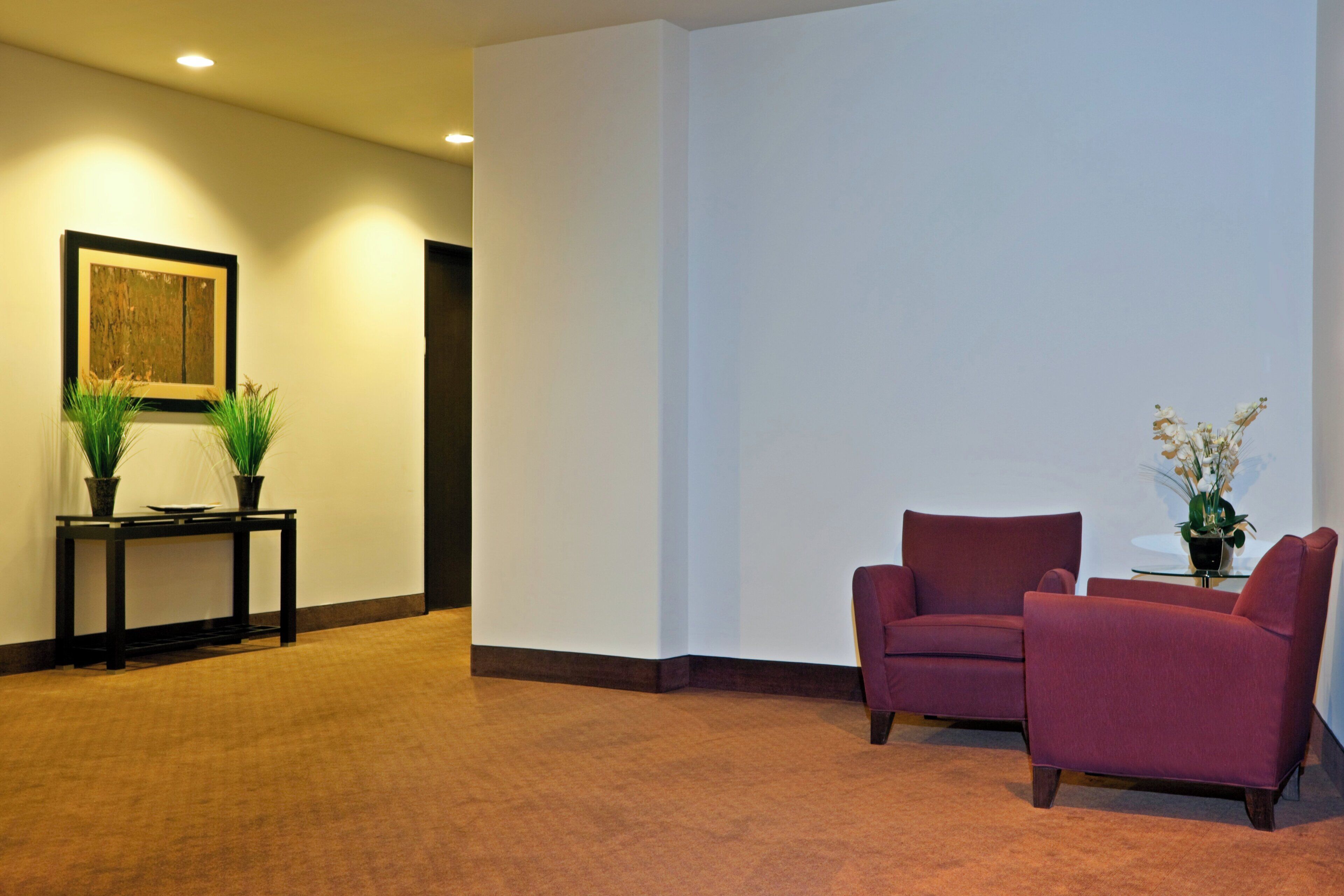 A hallway with a carpeted floor, a framed artwork on the wall, a small table with a plant, and two chairs.