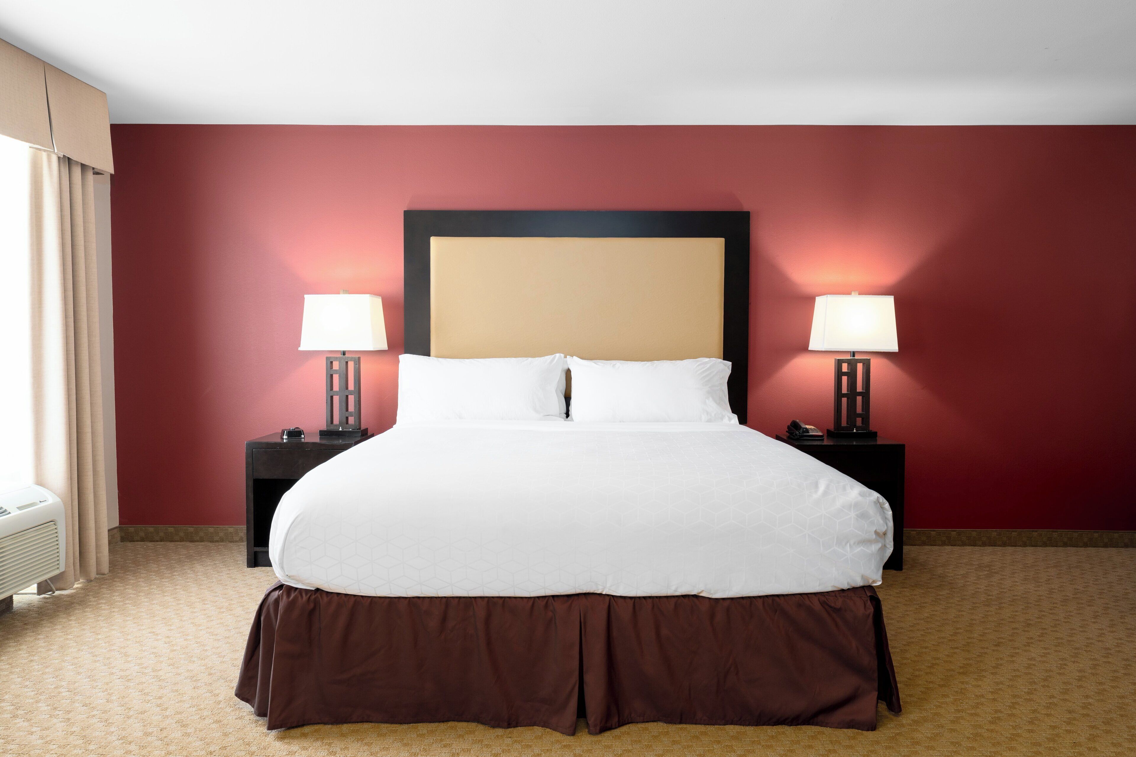 Une chambre d’hôtel avec un grand lit, deux tables de chevet avec des lampes, un mur rouge et une fenêtre avec des rideaux.