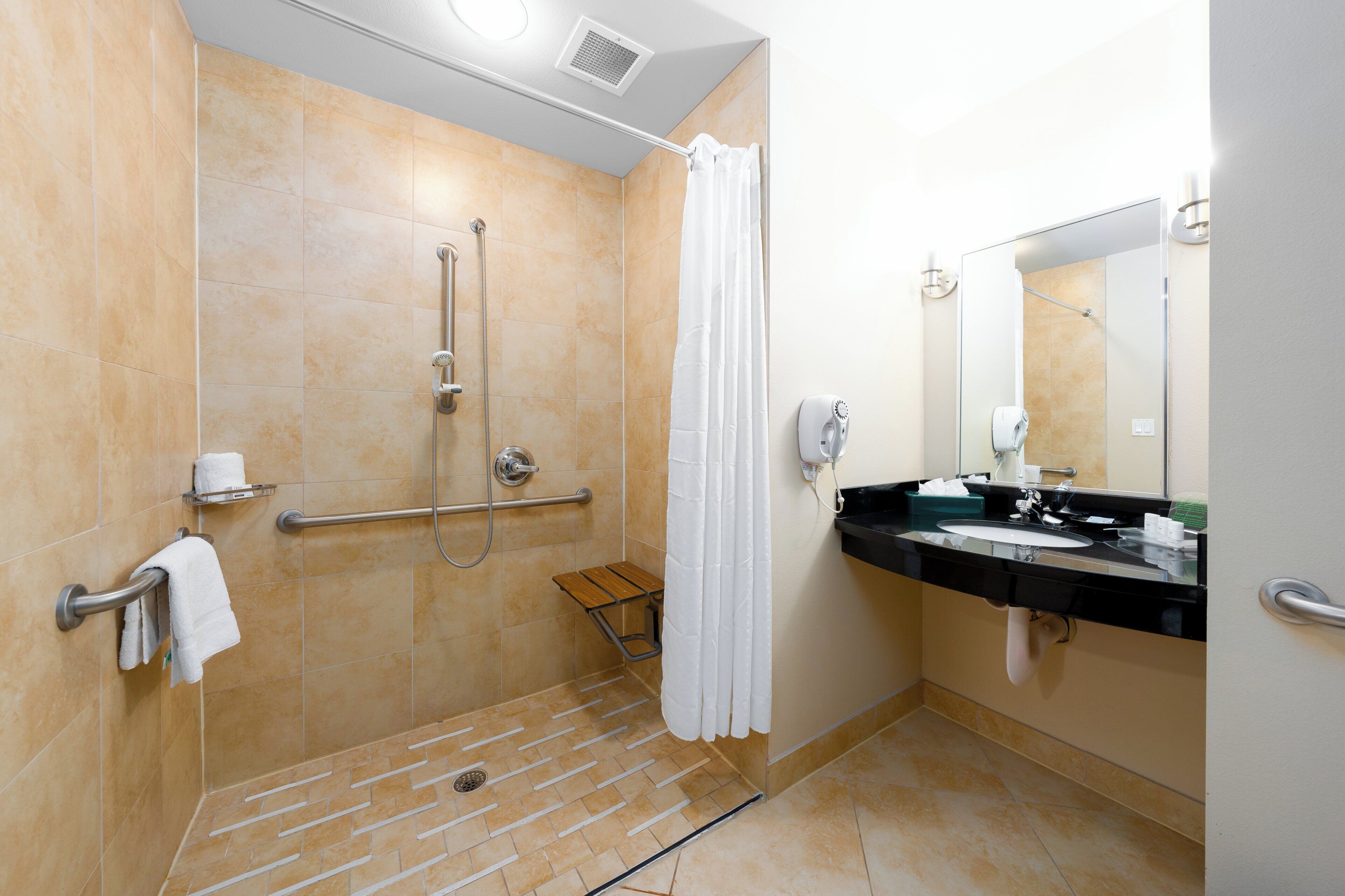 A bathroom with a walk-in shower, grab bars, a bench, a sink with a mirror, and a towel rack.