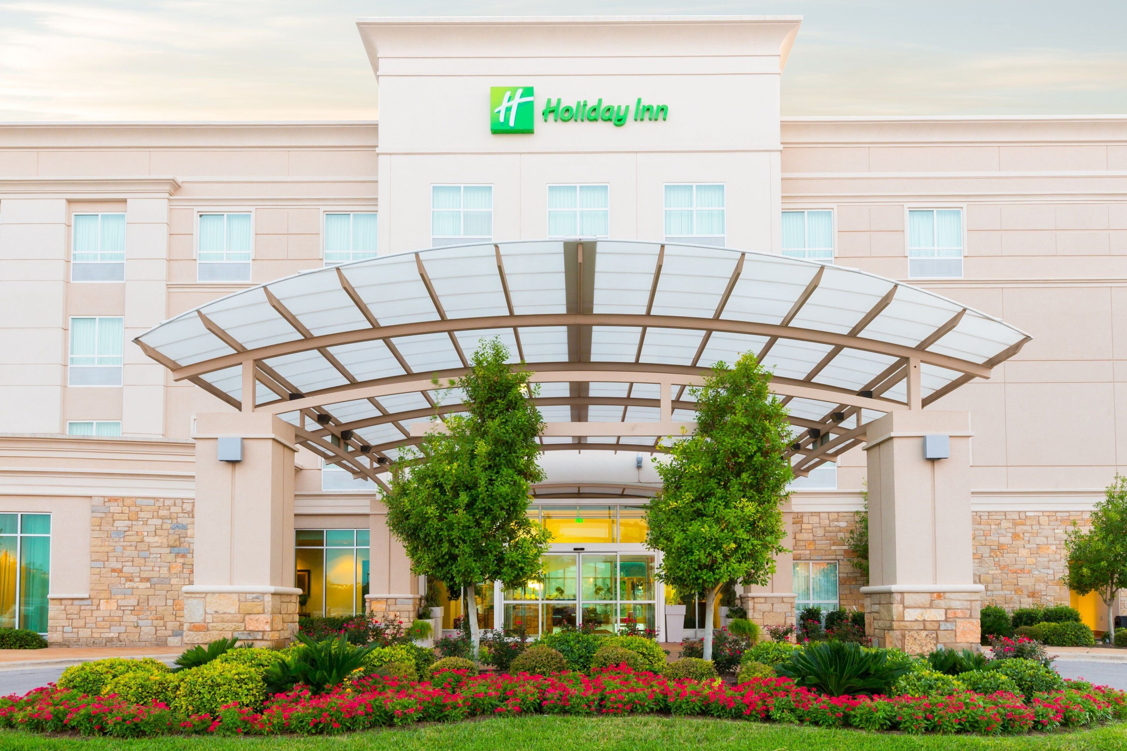 A Holiday Inn hotel with a covered entrance, multiple windows, and a landscaped garden with various plants and flowers.