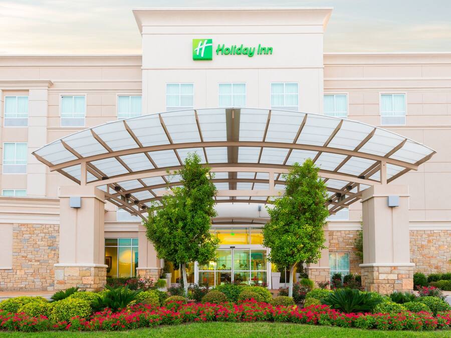 Un hotel Holiday Inn con entrada cubierta, múltiples ventanas y un jardín ajardinado con diversas plantas y flores.