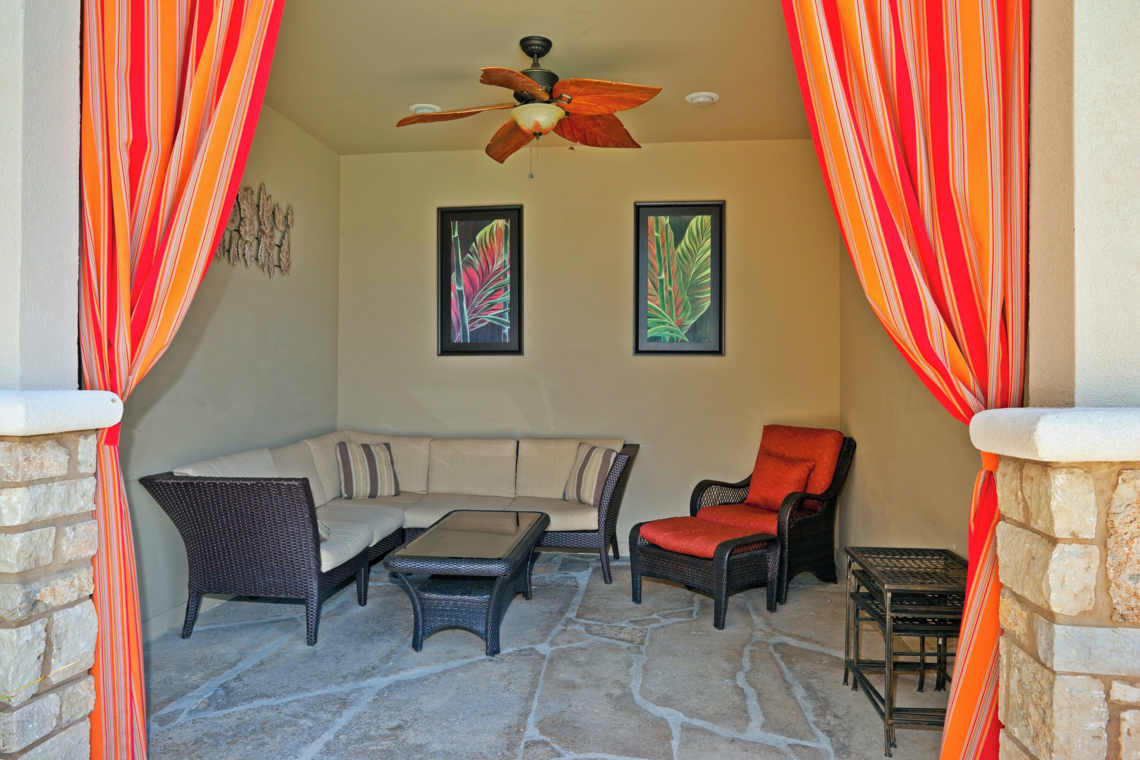A covered patio area with a stone floor, a sofa, two chairs, a table, and a ceiling fan.