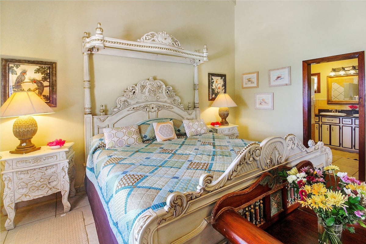 Uma cama com dossel e cabeceira decorada, duas mesas de cabeceira com abajures e um vaso de flores.