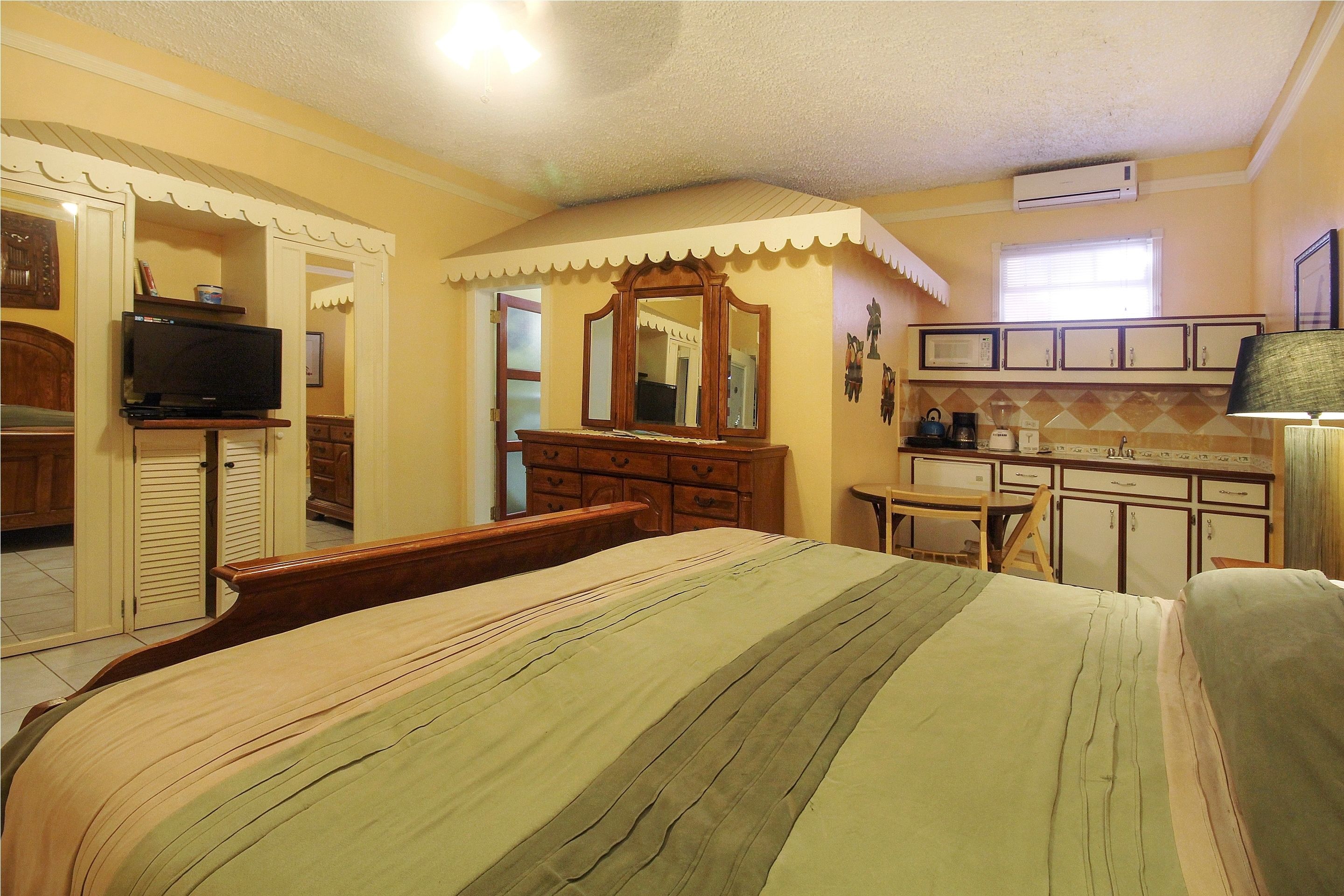 Un dormitorio con cama, cómoda, televisor y una kitchenette con armarios y microondas.
