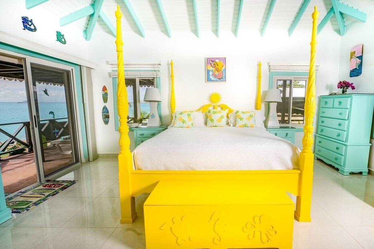 Um quarto claro com uma cama de dossel amarela, mobiliário azul-turquesa e vista para o mar através de uma porta deslizante.