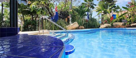 A swimming pool with a blue tiled deck, surrounded by tropical plants and palm trees.