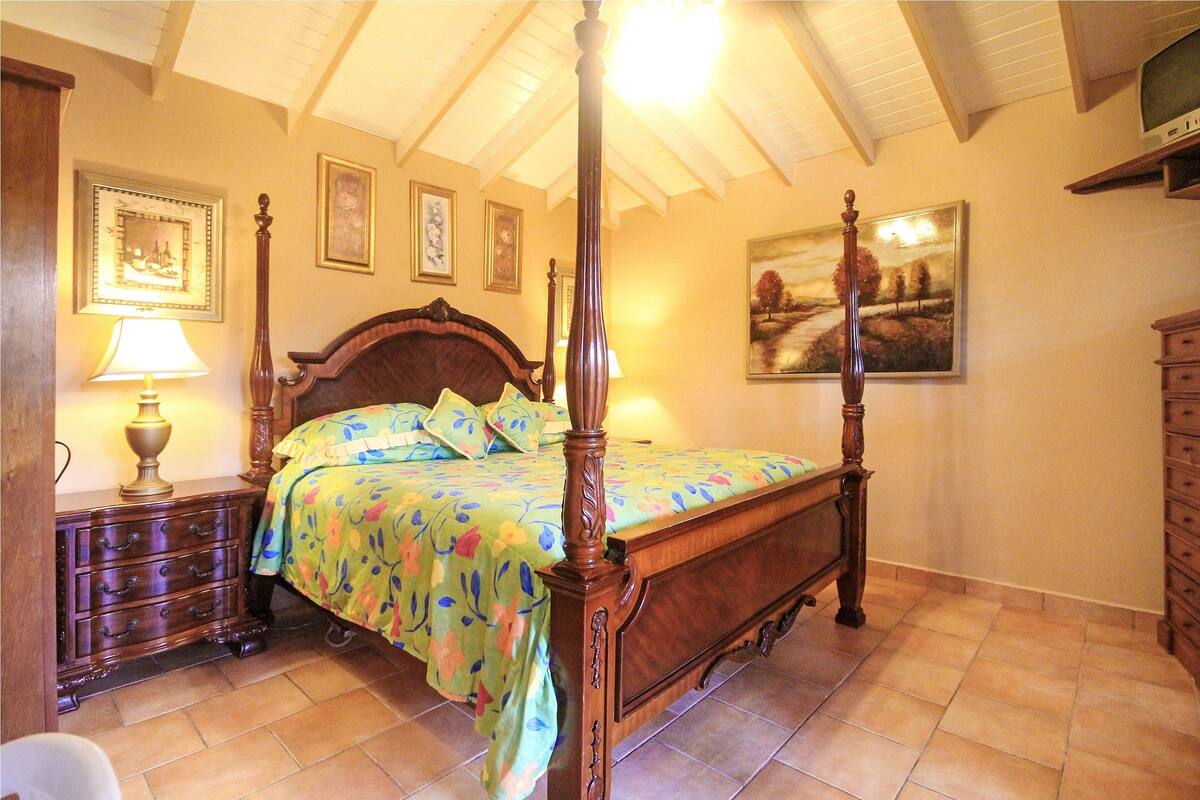 Uma cama com dossel adornada com roupa de cama florida, uma mesa de cabeceira de madeira com abajur e uma cômoda com gavetas.