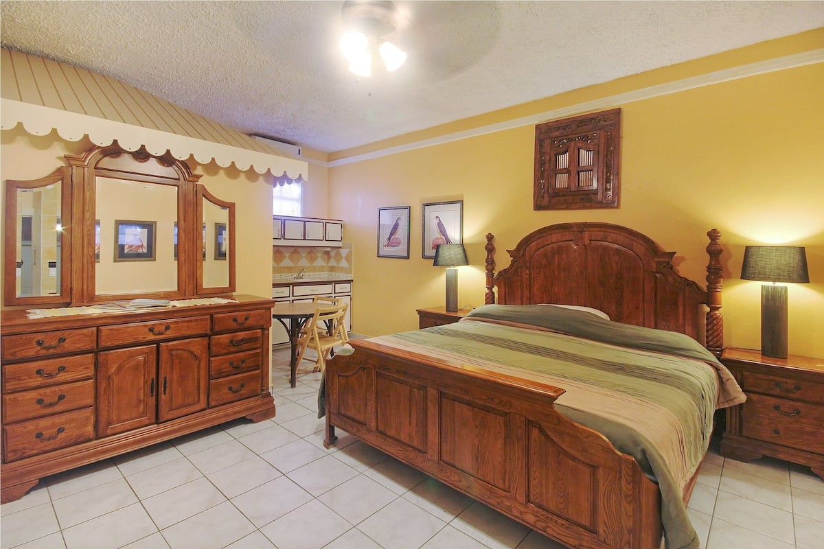 Quarto com cama de madeira, cômoda, penteadeira com espelho e escrivaninha com cadeiras.