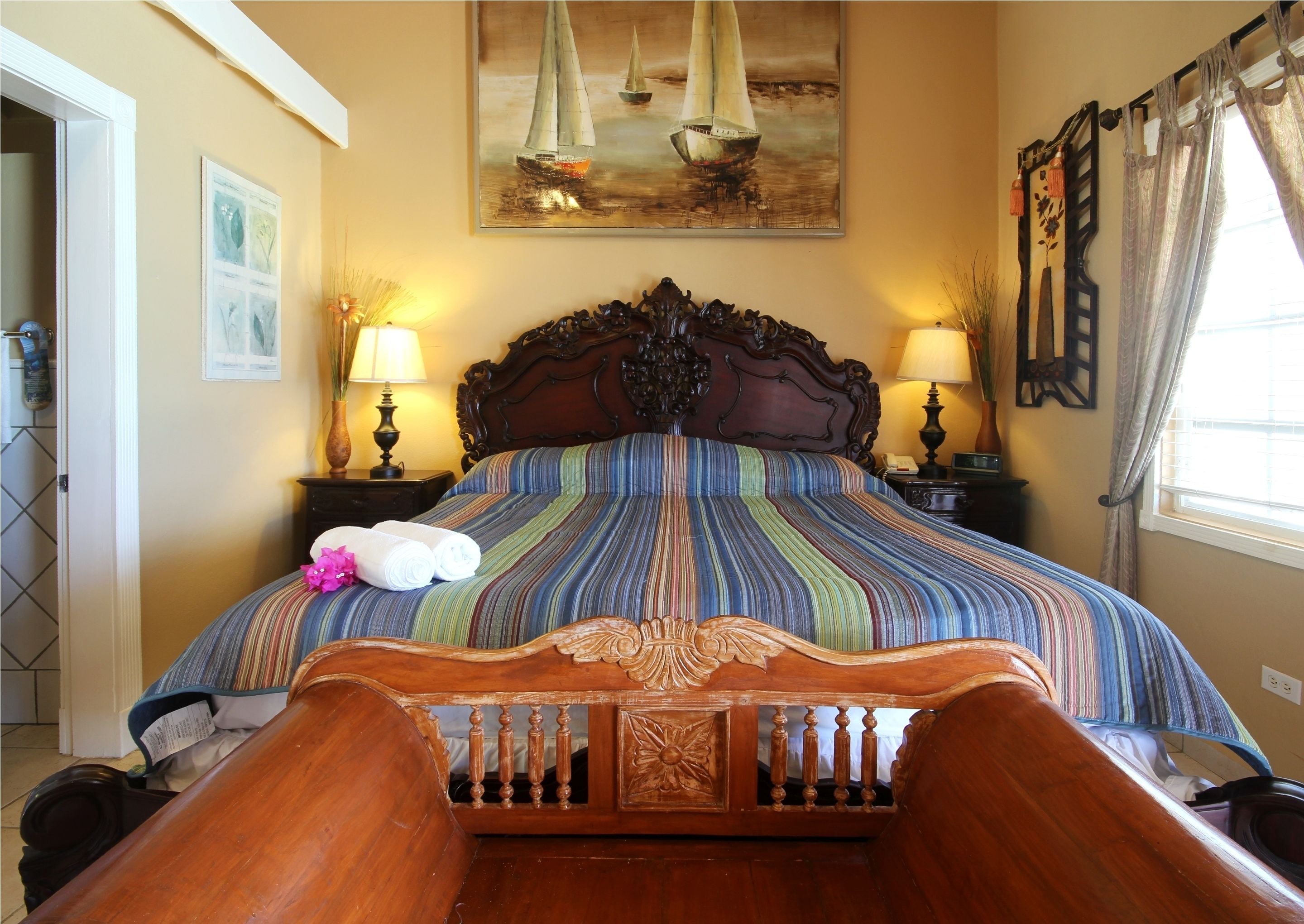 A bed with a striped bedspread, two bedside lamps, a painting of sailboats, and a wooden headboard.