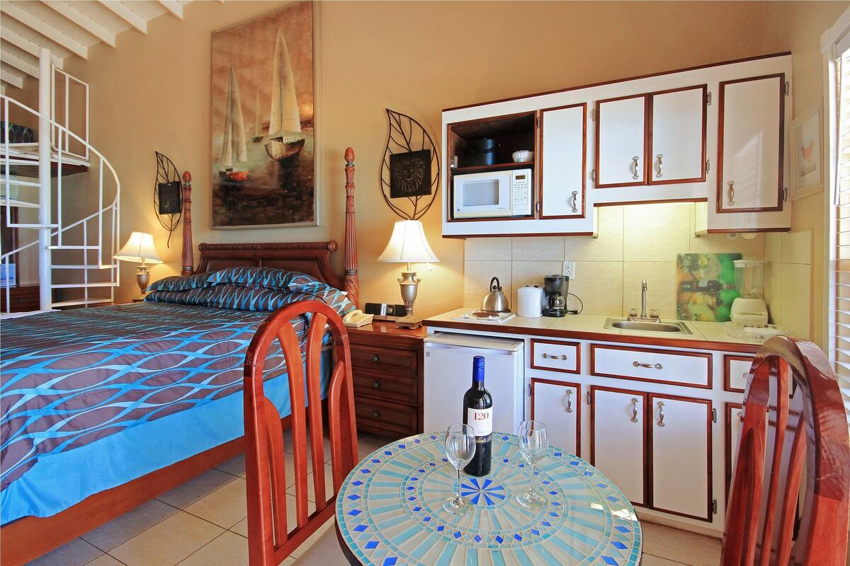 Quarto com cama, uma cadeira, mesa com garrafa e copos, escrivaninha com abajur e cozinha compacta com armários e micro-ondas.